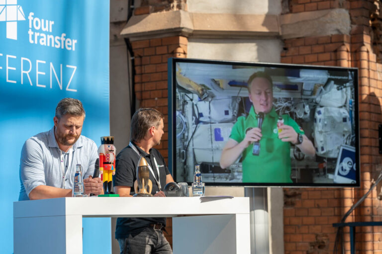 2 Männer halten einen Vortrag auf einer Bühne; auf einem Bildschirm ist ein Astronaut zu sehen