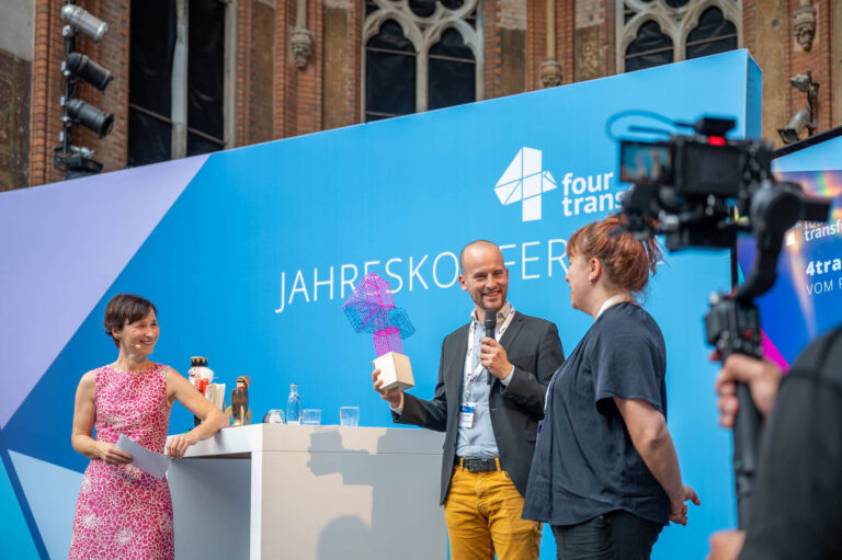 2 FRauen und ein Mann stehen auf einer Bühne und sprechen in Mirkofone; Der man hält eine Skulptur aus Kunststoff in der Hand