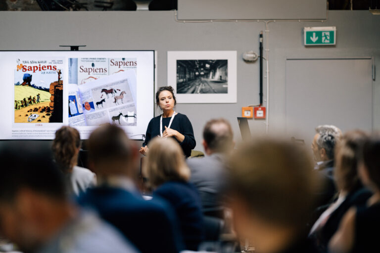 Eine FRau spricht vor Publikum in einem Workshop vor einem Board auf dem Ideen gesammelt werden